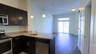 This single-floor space showcases an open-concept layout that seamlessly connects the kitchen to the living area, creating a cohesive and functional environment. The kitchen is appointed with dark espresso cabinetry that contrasts elegantly against light granite countertops, which extend along a peninsula-style counter. A stainless steel undermount sink and dishwasher add a modern, polished touch to the kitchen's design. Two pendant lights hang above the peninsula, providing focused task lighting while contributing to the overall aesthetic. The rich, dark hardwood flooring runs throughout the space, lending warmth and continuity between the kitchen and living areas. The living area benefits from abundant natural light streaming through a large sliding glass door, which opens to an outdoor balcony, enhancing the sense of openness. Neutral-toned walls complement the design palette and allow for versatile interior styling. The elevated ceilings further amplify the spacious feel of the interior. Overall, the space strikes a balance between contemporary design and everyday practicality, offering a bright and well-appointed environment suitable for comfortable living.