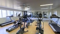 This single-level fitness center offers a thoughtfully arranged workout space designed to accommodate a variety of exercise routines. The room features light-toned hardwood-style flooring throughout most of the space, with a dark rubberized mat area designated for strength and weight training equipment. Natural light streams in through multiple windows along one wall, creating a bright and energizing atmosphere well-suited for exercise.

The cardio section is well-stocked with multiple elliptical trainers, stationary bikes, and additional aerobic machines positioned toward the front of the room. A multi-station cable and weight machine anchors the strength training zone toward the rear, alongside additional resistance equipment. Full-length mirrors line one wall, enhancing the sense of openness and allowing users to monitor their form during workouts.

Ceiling fans are installed throughout the space, providing consistent airflow and comfort during intense sessions. Recessed and linear fluorescent lighting supplements the natural light, ensuring the room remains well-lit at all hours. The overall layout is open and functional, offering a clean, organized environment suitable for a range of fitness activities.