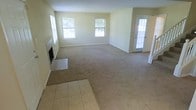 This 2-floor home features an open and generously sized main living area that serves as the centerpiece of the ground floor. The living room is finished with wall-to-wall carpet and neutral beige walls, creating a clean, understated aesthetic. A fireplace with a tiled hearth anchors one wall, adding a functional and visual focal point to the room. The tiled entryway near the fireplace transitions seamlessly into the carpeted living space, offering a practical and attractive flooring combination. Multiple windows along the far wall allow natural light to fill the room, contributing to a bright and airy atmosphere. A glass-paned exterior door provides additional light and access to the outdoors. The open layout of the main floor allows for flexible furniture arrangements and a natural flow between spaces. The overall design is straightforward and functional, with a neutral color palette that offers a versatile backdrop for a variety of interior styles. The second floor adds additional living space, expanding the home's overall capacity and utility.