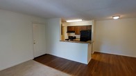 This single-floor residence offers a practical and functional layout designed to maximize the use of space. The open-concept living area features a combination of hardwood-style flooring and carpeting, creating a warm and inviting atmosphere throughout. A raised breakfast bar separates the kitchen from the living space, maintaining an open sightline while providing a natural division between the two areas. The kitchen is equipped with dark wood cabinetry, a black refrigerator, a gas range with an overhead exhaust hood, and a fluorescent ceiling light that brightens the cooking area. The countertops feature a speckled stone-style finish that complements the cabinetry. The entry door, positioned adjacent to the kitchen pass-through, features a classic white finish with gold-tone hardware, offering a clean and traditional look. The textured ceiling adds a subtle architectural detail consistent throughout the space. Overall, the unit presents a straightforward and functional design, well-suited for everyday living with its efficient use of layout and durable finishes throughout.
