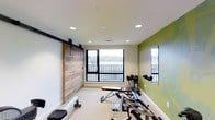 This single-floor fitness space is thoughtfully designed to inspire an active lifestyle, blending functional equipment with an inviting aesthetic. The room is anchored by a striking rustic barn door finished in reclaimed wood planking, which slides along a bold black track rail — a standout design element that adds warmth and character to the otherwise clean, modern interior. A vibrant olive-green accent wall provides an energizing pop of color, complemented by crisp white walls and a smooth white ceiling fitted with recessed lighting for bright, even illumination throughout. Large windows with black frames flood the space with natural light and offer pleasant outdoor views of surrounding greenery, creating an uplifting atmosphere during workouts. An HVAC vent ensures climate-controlled comfort year-round. The space is outfitted with a variety of strength and conditioning equipment, including a weight bench, dumbbells, and additional multi-use fitness machines, providing options for a well-rounded workout routine. The light-colored flooring is easy to maintain and keeps the space feeling open and airy. Whether used for strength training, stretching, or cardio work, this fitness room delivers a clean, motivating environment designed with both form and function in mind.