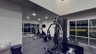 This well-appointed fitness center offers a thoughtfully designed workout environment suited for a variety of exercise routines. The space is characterized by impressively high ceilings that create an open, airy atmosphere, while large windows along the walls invite natural light and provide views of the surrounding outdoors, enhancing the overall workout experience.

The gym is equipped with a strong selection of cardio machines, including ellipticals and a stationary bike, positioned to take advantage of the open layout. A multi-station strength training machine anchors the space, offering users a versatile range of resistance exercises. Wall-mounted flat-screen monitors are conveniently positioned throughout, allowing users to stay entertained or track workout metrics during their sessions.

The flooring consists of a dark, rubberized material that provides both safety and durability, ideal for high-impact activities and heavy equipment. The neutral gray and white color palette of the walls contributes to a clean, modern aesthetic. Recessed lighting supplements the natural light, ensuring the space remains well-lit during evening hours. A mirrored wall section expands the visual depth of the room while allowing users to monitor their form. Overall, this fitness center delivers a functional, contemporary space designed to support a wide range of fitness goals.