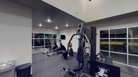 This well-appointed fitness center offers a thoughtfully designed workout environment suited for a variety of exercise routines. The space is characterized by impressively high ceilings that create an open, airy atmosphere, while large windows along the walls invite natural light and provide views of the surrounding outdoors, enhancing the overall workout experience.

The gym is equipped with a strong selection of cardio machines, including ellipticals and a stationary bike, positioned to take advantage of the open layout. A multi-station strength training machine anchors the space, offering users a versatile range of resistance exercises. Wall-mounted flat-screen monitors are conveniently positioned throughout, allowing users to stay entertained or track workout metrics during their sessions.

The flooring consists of a dark, rubberized material that provides both safety and durability, ideal for high-impact activities and heavy equipment. The neutral gray and white color palette of the walls contributes to a clean, modern aesthetic. Recessed lighting supplements the natural light, ensuring the space remains well-lit during evening hours. A mirrored wall section expands the visual depth of the room while allowing users to monitor their form. Overall, this fitness center delivers a functional, contemporary space designed to support a wide range of fitness goals.