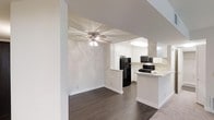 This single-floor residence offers a clean, open-concept design that connects the living area and kitchen in a cohesive, functional layout. The living space is finished with rich, dark hardwood flooring that extends throughout, providing a warm contrast against the bright white walls and trim. A ceiling fan with integrated lighting anchors the living area, contributing both style and comfort to the space.

The kitchen is thoughtfully designed with white shaker-style cabinetry that extends to the upper walls, maximizing storage while maintaining a bright, airy aesthetic. A peninsula counter with a light-toned surface serves as a natural divider between the kitchen and living areas, offering additional prep space and casual seating potential. Black appliances, including a refrigerator, gas range, and over-the-range microwave, provide a bold contrast to the white cabinetry and create a modern, high-contrast look.

The open sightlines between the two spaces make the floor plan feel expansive and well-connected, ideal for everyday living and entertaining. Neutral wall tones throughout enhance the sense of light and space, while clean white baseboards and architectural columns add subtle structural definition. The overall ambiance is bright, modern, and move-in ready.