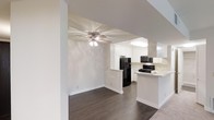 This single-floor residence offers a clean, open-concept design that connects the living area and kitchen in a cohesive, functional layout. The living space is finished with rich, dark hardwood flooring that extends throughout, providing a warm contrast against the bright white walls and trim. A ceiling fan with integrated lighting anchors the living area, contributing both style and comfort to the space.

The kitchen is thoughtfully designed with white shaker-style cabinetry that extends to the upper walls, maximizing storage while maintaining a bright, airy aesthetic. A peninsula counter with a light-toned surface serves as a natural divider between the kitchen and living areas, offering additional prep space and casual seating potential. Black appliances, including a refrigerator, gas range, and over-the-range microwave, provide a bold contrast to the white cabinetry and create a modern, high-contrast look.

The open sightlines between the two spaces make the floor plan feel expansive and well-connected, ideal for everyday living and entertaining. Neutral wall tones throughout enhance the sense of light and space, while clean white baseboards and architectural columns add subtle structural definition. The overall ambiance is bright, modern, and move-in ready.