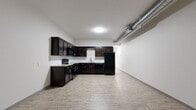 Located at 429 W Central Ave in Wichita, Kansas, this single-floor unit offers a thoughtfully designed interior with a clean, modern aesthetic throughout. The kitchen is a standout feature, outfitted with rich dark espresso cabinetry, granite countertops, a subway tile backsplash, and a full suite of black appliances including a range, over-the-range microwave, dishwasher, and refrigerator. Light wood-look flooring runs through the kitchen and open living areas, complementing the crisp white walls and creating a bright, airy atmosphere. Exposed ductwork adds an industrial character to the space, contributing to its urban loft sensibility. The bedroom is carpeted for comfort and features dual closet units with ample storage, along with a window overlooking mature trees that brings in natural light and greenery. A separate room with wood-look flooring and a panel entry door provides additional flexible living space. The bathroom includes a glass-enclosed shower with chrome fixtures. The overall floor plan, as revealed in the three-dimensional model, follows a rectangular layout with clearly defined zones for living, sleeping, and cooking, making efficient use of the available space throughout.