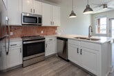 Located at 215 Willow Ave in Knoxville, Tennessee, this single-floor unit offers a clean, modern aesthetic with an open and airy layout. Light hardwood flooring flows seamlessly throughout the space, complementing the bright white walls and creating a cohesive, contemporary feel.

The kitchen is a standout feature, designed with white shaker-style cabinetry accented by matte black hardware and topped with light quartz countertops. A peninsula island with a double undermount sink and pendant lighting serves as a natural divider between the kitchen and living area. Stainless steel appliances — including a refrigerator, range, built-in microwave, and dishwasher — are paired with a distinctive terracotta-toned tile backsplash that adds a warm design accent.

The open living area extends naturally from the kitchen, with a ceiling fan overhead and access to an outdoor balcony through a glass door, allowing for ample natural light. The bedroom is generously proportioned, also featuring a ceiling fan and a private door leading to an outdoor area with a window view.

The bathroom offers a functional and stylish design, complete with a wide dual-sink vanity, quartz countertops, a large mirror with strip lighting, and a tiled walk-in shower. The overall layout of the unit prioritizes flow, practicality, and modern finishes throughout.