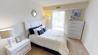 The bedroom is a thoughtfully designed space that balances comfort and style through a cohesive neutral palette. The room centers around a generously sized bed dressed in crisp white bedding and accented with navy blue and striped throw pillows, lending a fresh, classic feel. A tall white paneled headboard serves as a focal point, complementing the white nightstand and matching chest of drawers that provide ample storage. Light-toned carpeting adds warmth underfoot and contributes to the room's soft, restful ambiance. A large window dressed with horizontal blinds allows natural light to filter in, brightening the space and offering a view of the outdoors. A tripod floor lamp positioned near the window supplements the natural light, ensuring the room remains well-lit at any time of day. Decorative touches, including a vintage-style ornate mirror on the wall and a floral canvas print near the dresser, add personality without overwhelming the space. The neutral beige walls and visible ceiling vent suggest a practical, well-ventilated layout. Overall, the room presents a clean, serene environment well-suited for rest and relaxation.