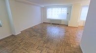 This single-floor space offers a generously proportioned open room defined by its classic parquet hardwood flooring, laid in a traditional block pattern that adds warmth and visual texture underfoot. The floors are well-maintained and catch natural light beautifully, highlighting their warm wood tones. A large window positioned along the far wall serves as a central architectural feature, flooding the interior with abundant natural light and offering views of the surrounding urban exterior. Beneath the window, a baseboard heating unit provides functional climate control. The walls are finished in a clean, neutral white throughout, creating a bright and versatile backdrop that complements the natural tones of the flooring. The ceiling features subtle detailing near the window wall, adding a modest architectural touch. The overall ambiance of the space is clean, airy, and open, with a classic character rooted in its traditional flooring design. The layout is straightforward and flexible, making it well-suited for a variety of uses. The combination of natural light, warm flooring, and neutral finishes gives the space a timeless, welcoming quality.
