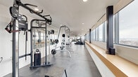 Located at 333 University Avenue in Kingston, Ontario, this contemporary fitness facility offers a thoughtfully designed, single-floor space that combines functionality with a clean, modern aesthetic. The layout features two distinct areas: a spacious main gym floor and an enclosed studio or flex room separated by full-height glass sliding doors, allowing for visual openness while maintaining acoustic separation.

The main gym area is generously proportioned, laid out in a long, corridor-style configuration that maximizes equipment placement without feeling congested. Large windows run continuously along one side, flooding the space with natural light and offering expansive elevated views of the Kingston skyline and surrounding landscape. A built-in wood-toned ledge beneath the windows provides a practical surface for accessories. The polished concrete-style tile flooring throughout lends the space a sleek, durable finish. Equipment includes cable and weight machines, treadmills, ellipticals, adjustable benches, plyo boxes, and free weights, with large wall mirrors enhancing the sense of depth.

The enclosed glass-walled room offers a quieter environment, featuring carpet flooring, a wall-mounted display, and the same panoramic views. Recessed lighting throughout ensures consistent illumination, while the crisp white walls and minimal design maintain a professional and welcoming atmosphere.