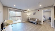 This thoughtfully designed one-bedroom, two-bathroom unit offers a clean, modern aesthetic across a single floor. The open-concept living area features light gray walls, recessed lighting, and light-toned hardwood-style flooring that runs seamlessly throughout the space. Large sliding glass doors lead to a private balcony that overlooks lush, wooded scenery, filling the living area with natural light and providing a serene outdoor connection. A wall-mounted mini-split HVAC unit ensures year-round climate control. The kitchen is efficiently integrated into the living space, featuring white cabinetry, granite-look countertops, a stainless steel refrigerator, and a peninsula that separates it from the main living area. The bedroom is a well-proportioned retreat with a dedicated window framing outdoor greenery, a wall-mounted mini-split unit, and recessed lighting. It connects to a bathroom equipped with a white vanity topped with granite counters, a walk-in closet with built-in shelving, and a tiled glass-enclosed stand-up shower. A second bathroom mirrors the clean, contemporary design. The overall ambiance is bright, neutral, and move-in ready, offering a functional and comfortable living environment.