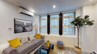 This single-floor residence is thoughtfully designed with a clean, modern aesthetic carried consistently throughout. The living area serves as a welcoming focal point, featuring a bank of large windows with bold black-framed detailing that floods the room with natural light and offers views of the surrounding cityscape. Light wood flooring, recessed lighting, a wall-mounted split AC unit, and curated décor accented in yellow and grey tones contribute to a polished, contemporary feel.

The kitchen is efficiently laid out with warm wood cabinetry extending to the ceiling, white quartz countertops, a white subway tile backsplash, and stainless steel appliances including a full-size refrigerator. A dining table sits adjacent, making the space both functional and inviting.

Two bedrooms provide comfortable accommodations. The primary bedroom benefits from a large window with urban views, a wall-mounted AC unit, and direct sightlines to a living area beyond. The second bedroom is quietly styled with framed artwork and a side window for additional natural light.

The bathroom is a standout, featuring patterned hexagonal mosaic floor tiles, a walk-in shower enclosed in black-framed glass with white subway tile walls, and warm wood vanity cabinetry.
