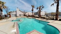 This residential community showcases a standout outdoor amenity area centered around a generously sized swimming pool with sparkling turquoise water. The pool's freeform design is complemented by a connected hot tub, brick-trimmed edges, and multiple entry points with stainless steel handrails for easy access. Tall palm trees flank the pool deck, lending a resort-like ambiance to the setting. The surrounding patio features open lounging areas shaded by a pergola-style structure, ideal for relaxing outdoors. A charming single-story clubhouse or amenity building with large French doors, a pitched roof, and a chimney sits directly adjacent to the pool, offering a seamless indoor-outdoor connection. The space is enclosed by a decorative iron fence, providing both privacy and a sense of security. Multi-story residential buildings are visible in the background, set among mature trees, giving the community a well-established feel. The open sky and abundant natural light enhance the inviting, warm atmosphere of this outdoor gathering space, making it a central social hub for residents throughout the warmer months.