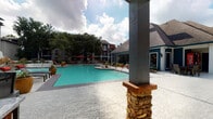 This outdoor amenity space is designed to offer a resort-style experience centered around a generously sized swimming pool with vivid turquoise water. The pool deck is finished in decorative concrete and features a shallow entry point, lounge seating, and shaded umbrella stations, all framed by mature trees and lush landscaping. Stone-accented columns define the perimeter of the space, adding architectural character to the open-air environment.

Adjacent to the pool is a covered outdoor kitchen pavilion, thoughtfully designed for communal entertaining. The pavilion features dual stainless steel built-in grills, extensive granite countertops, a utility sink, and cabinetry finished in a slate-gray beadboard style. Bar-height seating along the granite-topped counter accommodates multiple guests, making it an ideal setting for gatherings. The covered ceiling overhead provides shade and shelter, while open archways maintain a seamless connection to the surrounding pool area and greenery.

The overall ambiance of the space balances functionality with aesthetic appeal, offering a comfortable and attractive outdoor retreat. The combination of the shimmering pool, well-equipped outdoor kitchen, and carefully maintained landscaping creates a cohesive and inviting communal environment suited for both relaxation and social activities.