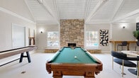 Located at 5242 Edmondson Pike in Nashville, TN, this single-floor game room is a well-appointed recreational space designed for both entertainment and relaxation. The room's most striking architectural feature is its dramatic vaulted ceiling with exposed white-painted wood beams and shiplap paneling, which creates an airy, open atmosphere and a sense of generous vertical space.

A floor-to-ceiling stacked stone fireplace serves as the room's focal point, flanked by large windows that invite abundant natural light and outdoor views. The fireplace's warm, earthy tones contrast beautifully with the crisp white ceiling and neutral walls, lending the space a refined yet inviting character.

The game room is well-equipped for entertainment, featuring a classic billiards table with a green felt surface and ornate carved wood legs, alongside a shuffleboard table for additional play. A stylish lounge area with a marble-top pedestal table and plush curved seating chairs provides a comfortable spot to unwind, while a bar-height counter with bar stools along one wall offers a dedicated social gathering area.

Contemporary pendant light fixtures, curated wall art, and a textured accent wall add sophisticated finishing touches. French doors provide access to the exterior, further enhancing the bright and welcoming ambiance of this thoughtfully designed recreational space.