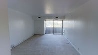 This single-floor space offers a straightforward and functional layout designed for comfortable everyday living. The main living area is finished with wall-to-wall carpeting that adds a soft, understated feel underfoot. Clean white walls and a smooth ceiling create a neutral backdrop that is easy to personalize and furnish to a variety of tastes.

A standout feature of the living area is the full-width sliding glass door fitted with vertical blinds, which opens directly onto an outdoor balcony. This connection to the outdoors allows for generous natural light to fill the room throughout the day while also providing treetop views that bring a sense of greenery and calm to the interior. The balcony itself extends the usable space and offers a pleasant spot for outdoor relaxation.

Ceiling-mounted ventilation registers indicate a central HVAC system, ensuring consistent climate control throughout the space. A single interior door provides access to additional areas of the unit. The overall design is clean, open, and efficient, with white walls and minimal ornamentation lending the space a bright, airy ambiance. The combination of natural light, outdoor access, and a neutral palette makes this a versatile space well-suited for a range of uses and personal styles.