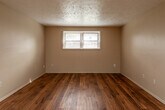 This single-story space offers a practical and straightforward layout across several rooms, each finished with consistent neutral tones and textured ceilings throughout. The main living area is generously sized, featuring warm hardwood flooring that extends into the kitchen and bathroom, providing a cohesive look across the common spaces. Natural light enters through blinded windows, giving the room an open, airy feel. A door provides direct access to an exterior area.

The kitchen is equipped with white cabinetry, dark countertops, a double-basin sink, and white appliances including a range, oven, and dishwasher. A sliding vertical blind door leads to an outdoor space, allowing additional natural light into the kitchen.

Two carpeted bedrooms each feature ceiling fans with integrated lighting, neutral wall tones, and windows with blinds. The carpet is plush and well-maintained, contributing to a comfortable atmosphere in each room.

The bathroom is finished with classic white tile accents featuring subtle green inlay details, hardwood flooring, a standard tub and shower combination, and a window for natural ventilation. A vanity with a dark countertop completes the space. Overall, the layout is compact, functional, and move-in ready.