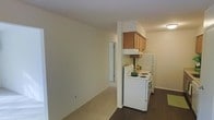 This single-floor residence offers a practical and straightforward layout designed for comfortable everyday living. The kitchen is efficiently arranged and features warm-toned wood upper cabinets mounted above a white electric range, providing a classic and clean aesthetic. A full-size refrigerator sits adjacent to the stove, maximizing the utility of the kitchen space. The flooring transitions from dark hardwood in the kitchen area to light neutral carpeting in the adjoining living space, creating a clear visual distinction between functional and relaxation zones. The living area is bright and open, with neutral white walls that lend a clean, versatile backdrop suitable for a variety of interior styles. A closet with a white bi-fold door is visible along one wall, offering convenient built-in storage. The overall ambiance of the unit is understated and practical, with a simple, uncluttered design that emphasizes usability. Recessed and overhead lighting provides adequate illumination throughout. The space is well-suited for those seeking a no-frills, move-in-ready layout with clearly defined living and kitchen zones within a compact and efficient single-level footprint.