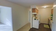 This single-floor residence offers a practical and straightforward layout designed for comfortable everyday living. The kitchen is efficiently arranged and features warm-toned wood upper cabinets mounted above a white electric range, providing a classic and clean aesthetic. A full-size refrigerator sits adjacent to the stove, maximizing the utility of the kitchen space. The flooring transitions from dark hardwood in the kitchen area to light neutral carpeting in the adjoining living space, creating a clear visual distinction between functional and relaxation zones. The living area is bright and open, with neutral white walls that lend a clean, versatile backdrop suitable for a variety of interior styles. A closet with a white bi-fold door is visible along one wall, offering convenient built-in storage. The overall ambiance of the unit is understated and practical, with a simple, uncluttered design that emphasizes usability. Recessed and overhead lighting provides adequate illumination throughout. The space is well-suited for those seeking a no-frills, move-in-ready layout with clearly defined living and kitchen zones within a compact and efficient single-level footprint.