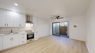 Located at 3265 C St in San Diego, CA, this efficiently designed single-floor unit offers a thoughtfully arranged layout comprising a living room, kitchen, bathroom, and hallway. The open-plan living and kitchen area is bright and modern, finished with light wood-toned flooring and crisp white walls that create a clean, airy atmosphere. A ceiling fan with integrated lighting anchors the living space, while a large sliding glass door floods the room with natural light and provides direct access to a private outdoor area enclosed by a wood-panel fence. The kitchen is outfitted with white flat-front cabinetry featuring matte black hardware, light stone countertops, a stainless steel undermount sink, a stainless steel refrigerator, and a gas range with a wall-mounted ventilation hood — delivering a fully functional and contemporary cooking space. The bathroom features consistent light wood flooring, a full tub-shower combination in a clean white surround, and a vanity with matching countertop. Throughout the unit, the consistent use of white tones, light flooring, and modern fixtures creates a cohesive, minimalist aesthetic well-suited to comfortable urban living in San Diego.