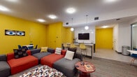 This lively media room is designed with a bold, modern aesthetic that immediately draws the eye. Striking yellow walls energize the space and provide a vivid contrast against crisp white surfaces and light tile flooring near the entrance. The lounge area is anchored by a generously sized sectional sofa in gray and red, complemented by a colorful hexagonal ottoman and a warm wood coffee table — all arranged around a wall-mounted flat-screen television. A standout feature is the graphic geometric mural wallpaper that adorns one wall, adding an artistic, modern touch to the TV viewing area. Adjacent to the lounge is a bar-height counter with granite-style surfaces and white subway tile backsplash, paired with a row of modern barstools — perfect for casual dining or socializing. A separate dining nook near the entry features round white tables and vibrant blue mid-century style chairs, offering additional seating options. The flooring transitions from large-format tile near the entry to a patterned carpet in the lounge area, clearly defining each zone. Recessed lighting along with globe pendant fixtures provide warm, even illumination throughout. Curated wall art and a small bookshelf add personality and a welcoming, communal feel to the overall space.