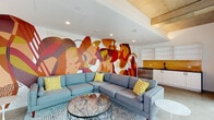 This single-floor amenity space is situated within a contemporary multi-story building with a striking modern exterior. The centerpiece of the interior is a sweeping, full-wall mural rendered in bold tones of red, orange, gold, and burgundy, filling the lounge area with vivid artistic energy. The lounge is furnished with a generously sized sectional sofa in cool grey upholstery, accented with yellow patterned throw pillows, and anchored by a round glass-top coffee table on a patterned area rug. Recessed lighting illuminates the space from above, while exposed concrete structural elements lend an industrial character to the ceiling. A wall-mounted flat-screen television provides an entertainment focal point opposite the seating arrangement. Adjacent to the lounge, a compact kitchenette features white cabinetry with open upper shelving, a dark countertop, a stainless steel undermount sink with a matte black faucet, and a vivid golden-yellow vertical tile backsplash that ties the color palette together. A beverage refrigerator completes the wet bar setup. The space also opens to a spacious outdoor courtyard deck with warm wood-toned decking, modular grey outdoor seating, a low coffee table, and accent chairs, all surrounded by young trees, planted greenery, and exposed concrete building facades — offering a tranquil urban retreat.