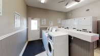 This shared laundry facility is designed to efficiently serve a residential community, offering a practical and organized environment for residents' laundering needs. The single-level space is outfitted with multiple top-load and front-load washing machines alongside a bank of stacked dryers, providing ample capacity for simultaneous use. A central folding counter runs through the space, offering a generous surface for sorting and folding laundry. The walls are finished in a neutral gray tone with a wainscoting-style lower panel, giving the room a clean and orderly appearance. Light-colored tile flooring keeps the space bright and easy to maintain. Overhead fluorescent lighting and a ceiling fan ensure the room stays well-lit and ventilated. An electronic payment kiosk is conveniently mounted on the wall, enabling cashless transactions. A cork bulletin board displays community notices, while instructional and service signage is posted throughout for user guidance. A vending machine provides added convenience for residents. A fire extinguisher is visibly mounted for safety. Natural light is introduced through a windowed entry door, complementing the artificial lighting. The facility is clearly maintained with user convenience and functionality as its core focus.