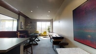 Located at 618 S First St in the heart of downtown San Jose, this single-floor social lounge is thoughtfully designed for recreation and community engagement. The space is anchored by a full-size foosball table and a long shuffleboard table, providing ample entertainment options for groups. Recessed lighting illuminates the patterned carpet flooring, while large floor-to-ceiling windows flood the room with natural light and offer open views of the surrounding urban streetscape.

A cozy lounge corner features a pair of bold, quilted golden-yellow accent chairs arranged around a small side table, creating an inviting nook for relaxed conversation. The walls are adorned with curated artwork, including abstract paintings and dimensional wall sculptures, adding a distinctive artistic character to the space. Deep burgundy booth-style seating along one wall provides additional gathering spots, complemented by high-top tables and bar seating.

A built-in kitchenette with dark cabinetry, a stainless steel undermount sink, and a quartz countertop serves as a practical refreshment station. The space carries a maximum occupancy of 32, making it well-suited for private events and social gatherings. The overall ambiance balances modern, contemporary design with a warm, welcoming atmosphere.