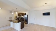 Located at 115 Amberleigh Dr in Wilmington, North Carolina, this single-floor residence offers a well-designed, open-concept layout that seamlessly connects the kitchen and living areas. The kitchen is a standout feature, showcasing rich dark wood cabinetry paired with dark granite countertops that extend to a functional breakfast bar island. Stainless steel appliances — including a refrigerator, built-in microwave, and range — lend a modern, polished look, while pendant lighting and a track light fixture above illuminate the space with warm, focused light. The kitchen flows naturally into the carpeted living area, which is bright and open with clean white walls that enhance the sense of spaciousness. Tile flooring in the kitchen and entryway transitions smoothly into plush carpet in the living space. The vaulted-style ceiling adds a sense of height and airiness throughout. A white interior door leads to additional areas of the home. The overall ambiance is clean, contemporary, and functional, making this residence a comfortable and practical living space in the Wilmington, NC area.