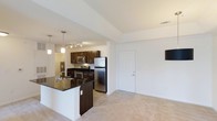 Located at 115 Amberleigh Dr in Wilmington, North Carolina, this single-floor residence offers a well-designed, open-concept layout that seamlessly connects the kitchen and living areas. The kitchen is a standout feature, showcasing rich dark wood cabinetry paired with dark granite countertops that extend to a functional breakfast bar island. Stainless steel appliances — including a refrigerator, built-in microwave, and range — lend a modern, polished look, while pendant lighting and a track light fixture above illuminate the space with warm, focused light. The kitchen flows naturally into the carpeted living area, which is bright and open with clean white walls that enhance the sense of spaciousness. Tile flooring in the kitchen and entryway transitions smoothly into plush carpet in the living space. The vaulted-style ceiling adds a sense of height and airiness throughout. A white interior door leads to additional areas of the home. The overall ambiance is clean, contemporary, and functional, making this residence a comfortable and practical living space in the Wilmington, NC area.