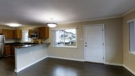 This single-floor space offers an efficient, open-concept layout that seamlessly connects the kitchen and main living area. The kitchen is equipped with warm honey-toned wood cabinetry, a stainless steel microwave, a gas range, and a dark granite or laminate countertop that extends into a peninsula-style breakfast bar, providing a natural division between the kitchen and the adjoining open area. Crown molding runs along the ceiling perimeter, adding a clean, polished finish to the interior. The flooring throughout is a rich, dark hardwood, lending warmth and cohesion to the space. A pendant light fixture with a frosted bowl shade hangs centrally, providing ambient illumination to the main area. A large window overlooks the front exterior, allowing natural light to filter in and offering views of the surrounding neighborhood. Adjacent to the window, a paneled front entry door with brushed hardware provides direct access to the outdoors. The textured ceiling and neutral wall tones contribute to a practical, understated aesthetic throughout the space.