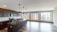 This single-floor residence showcases a thoughtfully designed open-concept layout that seamlessly connects the kitchen, dining, and living areas. The kitchen is a standout feature, boasting rich dark-wood cabinetry extending to the ceiling, dark granite countertops, a white subway tile backsplash, and a generously sized central island with an integrated sink. Stainless steel appliances, including a range, microwave, and refrigerator, complete the well-appointed culinary space. Three stylish pendant lights hang above the island, adding a modern aesthetic touch.

The expansive living area is bathed in natural light from large, industrial-style grid windows that frame views of the surrounding urban landscape. Durable hardwood flooring runs throughout the main living spaces, contributing warmth and continuity. The bathroom features neutral tile flooring, white cabinetry, white subway tile accents, and dark-finished panel doors that provide a cohesive design element across the unit. A spacious walk-in closet with built-in shelving and hanging rods offers substantial storage. The overall ambiance is contemporary and polished, balancing urban style with practical functionality.