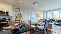 Located at 2 Center St in the heart of Newark, NJ, this thoughtfully designed single-floor residence offers a seamless blend of modern style and functional living. The open-plan layout connects the kitchen, dining, and living areas, creating a cohesive and inviting atmosphere throughout.

The kitchen is outfitted with stainless steel appliances, crisp white cabinetry, dark granite countertops, and rich hardwood flooring, providing both aesthetic appeal and practical utility. A tiered display stand and generous counter space make it ideal for cooking and entertaining.

The dining area features a glass-top round table beneath a striking cascading chandelier, adding an elegant focal point to the space. This flows naturally into the living room, where floor-to-ceiling windows frame impressive city views of the Newark skyline, filling the room with abundant natural light. A deep navy sectional sofa, patterned area rug, and curated artwork complete the refined aesthetic.

The bedroom offers a serene retreat with plush carpeting, ample closet space, and tasteful décor. The bathroom is finished with large-format tile, a soaking tub, dark granite vanity, and white cabinetry, delivering a clean and functional design. Overall, the space presents a polished urban living experience in a prime downtown Newark location.