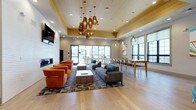 This contemporary communal space is thoughtfully designed to accommodate a variety of social and functional needs within a single open-plan floor. The main lounge area is anchored by a striking cluster of copper pendant lights suspended from a warm wood-paneled ceiling detail, creating a focal point that draws attention upward and adds visual warmth. Below, light hardwood flooring extends throughout, unifying the distinct zones seamlessly.

The lounge is organized into multiple seating arrangements, including a grouping of bold orange barrel chairs positioned around a slatted wood coffee table, facing a wall-mounted flat-screen television above a sleek linear gas fireplace set into a textured white brick feature wall. A second seating area features a charcoal sectional sofa accented with teal throw pillows, centered on a geometric patterned area rug.

Along the perimeter, a high-top bar counter with seating and pendant lighting provides additional workspace or casual dining. A dedicated dining table with chairs and vibrant abstract artwork lines the windowed wall, drawing in natural light and outdoor views. A compact kitchenette with wood cabinetry, dark granite countertops, and stainless steel appliances supports catering needs. Sliding glass doors connect the interior to an outdoor terrace.