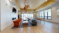 This contemporary communal space is thoughtfully designed to accommodate a variety of social and functional needs within a single open-plan floor. The main lounge area is anchored by a striking cluster of copper pendant lights suspended from a warm wood-paneled ceiling detail, creating a focal point that draws attention upward and adds visual warmth. Below, light hardwood flooring extends throughout, unifying the distinct zones seamlessly.

The lounge is organized into multiple seating arrangements, including a grouping of bold orange barrel chairs positioned around a slatted wood coffee table, facing a wall-mounted flat-screen television above a sleek linear gas fireplace set into a textured white brick feature wall. A second seating area features a charcoal sectional sofa accented with teal throw pillows, centered on a geometric patterned area rug.

Along the perimeter, a high-top bar counter with seating and pendant lighting provides additional workspace or casual dining. A dedicated dining table with chairs and vibrant abstract artwork lines the windowed wall, drawing in natural light and outdoor views. A compact kitchenette with wood cabinetry, dark granite countertops, and stainless steel appliances supports catering needs. Sliding glass doors connect the interior to an outdoor terrace.