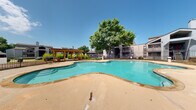 This outdoor amenity space centers around a spacious, freeform swimming pool with striking turquoise water, framed by a wide concrete pool deck ideal for lounging and recreation. The pool features clearly marked depth indicators and posted safety signage, reflecting a well-maintained and safety-conscious environment. A rustic wooden pergola on one side of the deck provides shaded seating, offering a comfortable retreat from the sun. Mature trees lend natural shade and a sense of established greenery to the setting, enhancing the overall ambiance of the space. The pool area is enclosed by a black wrought iron fence, providing both security and an open, airy aesthetic that allows unobstructed views of the surrounding landscape. A stacked stone retaining wall borders one edge of the property, adding a natural, textured architectural element that complements the outdoor setting. Lush green lawns and ornamental plantings are visible beyond the fence line, contributing to the verdant surroundings. The multi-story residential buildings adjacent to the pool feature a contemporary design with angular rooflines, balconies, and exterior staircases. Under clear blue skies, this pool area presents an inviting and functional outdoor retreat well-suited for warm-weather enjoyment.