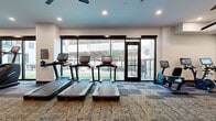 This expansive fitness center is thoughtfully designed to accommodate a full range of workout activities across its single-story layout. The main exercise floor is finished with durable patterned carpet tile and illuminated by recessed lighting and multiple ceiling fans, creating a comfortable environment for extended training sessions. Large floor-to-ceiling windows and glass doors line one side of the space, flooding the interior with natural light and offering views of an adjacent outdoor green area, lending the gym an open, airy feel.

The cardio zone features a generous row of treadmills equipped with individual touchscreen consoles, alongside elliptical trainers, stair climbers, and rowing machines. The strength training area is well-appointed with a full cable and multi-functional machine system, a comprehensive dumbbell rack displayed along a wall-length mirror, adjustable benches, medicine balls, and stability equipment. Ample floor space between stations ensures comfortable movement throughout.

A separate studio room provides a distinct environment for yoga, stretching, or group fitness, featuring light wood-look flooring, exposed wood beam ceilings, wood-panel accent walls, large mirrors, pendant lighting, and stability balls for flexible use.