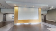 Located at 9502 Erickson Dr in Burnaby, BC, this residential lobby offers a welcoming and thoughtfully designed entrance experience. The generously proportioned hallway is finished with large-format tile flooring in a neutral grey palette, lending a clean and contemporary aesthetic throughout the space.

A striking central feature wall clad in warm amber wood paneling is paired with a textured, backlit white panel that serves as a visual focal point, adding depth and architectural interest to the interior. Recessed ceiling lighting complements the ambient illumination, creating a bright and inviting atmosphere.

Floor-to-ceiling glazing along one side of the lobby floods the space with natural light and frames lush outdoor greenery, bringing a sense of the outdoors inside. This windowed seating area is appointed with a sleek dark sofa and tall potted plants, creating a comfortable and verdant retreat for residents.

Along one wall, a refined vertical-ribbed panel treatment adds texture and visual rhythm, while a baseboard heating system ensures year-round comfort. Practical building amenities, including a mailbox station and access doors, are thoughtfully integrated without disrupting the lobby's polished appearance.

Overall, the space balances modern design sensibility with functional residential needs, offering residents a sophisticated and comfortable point of arrival.