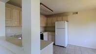 This two-floor space includes a kitchen designed for practical use. The kitchen features light-toned wood cabinetry that extends to the upper walls, providing ample storage throughout. A black gas range sits recessed within the lower cabinetry, complemented by a full-size white refrigerator positioned along the adjacent wall. The countertops are finished in a neutral tone, offering functional workspace, and a partial dividing wall creates a subtle separation between the kitchen and the neighboring area, lending a semi-open layout. Light tile flooring runs throughout, contributing to a clean, easy-to-maintain surface. Overhead, a rectangular fluorescent light fixture with wood trim detailing provides direct task lighting to the kitchen workspace. A visible ceiling vent indicates a central air system is in place. The neutral color palette of whites and beiges throughout the kitchen creates a straightforward, understated aesthetic. The two-floor layout of the overall space suggests additional living areas beyond what is visible, offering room for varied uses across both levels.