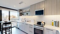 This well-appointed 1-bedroom, 1-bathroom residence offers a thoughtfully designed open-concept layout that maximizes every square foot. The kitchen and living area flow seamlessly together, anchored by a sleek, modern kitchen featuring light wood cabinetry, open display shelving with under-cabinet lighting, a white hexagonal tile backsplash, white quartz countertops, and a full suite of stainless steel appliances including a range, dishwasher, and built-in microwave. Large floor-to-ceiling windows flood the living area with natural light while framing impressive urban skyline views, creating an inviting and airy atmosphere. The living space comfortably accommodates a full sectional sofa and dining area. The bedroom is a comfortable and calm retreat with warm wood-tone flooring, soft neutral walls, and direct sightlines to the living area and city beyond. A dedicated walk-in closet provides ample storage with full-length hanging rods. The bathroom is finished in large-format white tile with a light wood vanity, sleek matte black hardware, and clean, minimalist design. Wood-look plank flooring runs throughout the main living spaces, tying the contemporary aesthetic together cohesively.