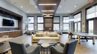 The community clubroom at Station Village at Avenel is a thoughtfully designed social hub that seamlessly blends comfort and functionality. The space is anchored by a striking floor-to-ceiling brick fireplace that serves as the room's focal point, flanked by large windows fitted with roller shades that flood the interior with natural light while maintaining a refined aesthetic. The ceiling design is a standout feature, combining recessed lighting with a bold wood-slat panel inset and suspended linear light fixtures that add warmth and visual depth overhead. The lounge area offers generous seating with a plush sofa, patterned accent pillows, and a pair of upholstered armchairs arranged around a central coffee table — ideal for relaxed conversation or casual gatherings. A ping-pong table positioned toward the rear of the room adds a recreational element, while high-top tables and stools line the perimeter near the windows, providing additional spaces for socializing or working. The hardwood-style flooring and neutral gray-and-gold color palette create a cohesive, contemporary atmosphere throughout. The open layout and abundance of natural light give the space an airy, welcoming character suitable for a wide range of community activities.