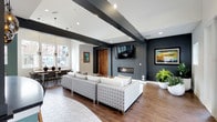 This contemporary single-floor space makes a strong first impression with its striking design elements and thoughtful layout. Exposed dark beams contrast sharply against a crisp white ceiling, creating a bold architectural statement that draws the eye throughout the room. Recessed lighting and pendant fixtures work together to provide layered illumination across the space.

The main lounge area is anchored by a generous sectional sofa upholstered in a geometric patterned fabric, positioned in front of a sleek linear fireplace set into a deep navy accent wall. A wall-mounted flat-screen television sits above, making this zone ideal for relaxation and entertainment. The flooring transitions thoughtfully between large hexagonal white tiles and rich wood-toned planks, subtly defining the different functional areas.

Adjacent to the lounge, a dining area benefits from natural light streaming through large windows that offer views of the surrounding streetscape. A warm wooden door adds a natural, organic touch amid the modern palette of dark navy, white, and charcoal tones. Lush potted greenery is incorporated throughout, softening the bold color contrasts and adding a welcoming, lively energy. Overall, the space delivers a refined yet comfortable ambiance suited for both social gatherings and quiet repose.
