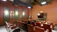 This private screening room delivers an authentic cinematic experience within a single-floor layout designed entirely for entertainment. The space features tiered, stadium-style seating with rows of rich leather chairs arranged to ensure unobstructed sightlines to the large flat-screen display mounted at the front of the room. The warm, terracotta-hued walls create a cozy, immersive atmosphere reminiscent of a classic movie house. Vintage-style film posters adorn the walls, adding a nostalgic and decorative touch that reinforces the cinematic theme. Soft wall sconces with green accents provide ambient lighting along the side walls, complementing the overhead industrial-style pendant lights that hang from dark, exposed ceiling beams. The ceiling's raw, structural aesthetic contrasts beautifully with the warmth of the interior, lending the room an inviting yet dramatic character. A built-in media cabinet or display case is visible toward the front of the room, offering additional storage or display functionality. The overall design balances comfort and style, making this space ideal for private film screenings, presentations, or group entertainment gatherings.