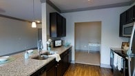 This single-floor residence presents a thoughtfully designed interior with a cohesive, contemporary aesthetic throughout. The kitchen serves as a central highlight, featuring rich dark wood cabinetry that extends to the upper walls, providing generous storage space. Granite countertops add a refined touch, complementing the stainless steel double-basin sink equipped with a modern arc faucet. Warm hardwood flooring runs through the main living areas, creating a sense of continuity and warmth. A pendant light fixture contributes focused illumination, enhancing the kitchen's functional appeal. The open sightlines connect the kitchen to an adjoining room, visible through a wide entryway, which features neutral carpeting and lighter wall tones — offering a calm, versatile space suitable for a variety of uses. A petite accent table tucked within this secondary room adds a charming, understated detail. The overall color palette of soft grays, deep espresso tones, and natural wood finishes creates a balanced and welcoming atmosphere. The layout maximizes the use of available space efficiently, making it a practical and attractive living environment that combines modern design sensibilities with everyday functionality.