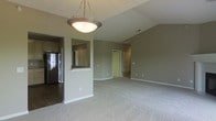 This single-floor space showcases an open-concept layout that seamlessly connects the kitchen and living areas. The living area is finished with plush neutral carpeting, while the kitchen features hard flooring, creating a natural visual distinction between the two spaces. Vaulted ceilings add a sense of height and openness to the main living area, enhancing the overall feeling of spaciousness.

A pass-through window with a countertop bar connects the kitchen to the living space, making it ideal for casual dining or entertaining. The kitchen is equipped with stainless steel appliances, including a refrigerator, visible from the pass-through. Walls are painted in a soft, warm gray-green tone throughout, contributing to a cohesive and calming aesthetic.

A pendant light fixture with a bowl-shaped shade hangs above, providing central illumination to the open area. Toward the back of the living space, a doorway leads to an additional room featuring louvered closet doors, suggesting a bedroom or utility space. White baseboards and trim provide clean contrast against the neutral walls, completing a well-maintained and functional interior.