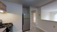 This single-floor space presents a clean, straightforward layout with a practical design. The kitchen area is equipped with a stainless steel top-freezer refrigerator, set against smooth white walls that contribute to a bright, neutral atmosphere throughout. The flooring features light-toned tiles, adding a subtle texture to the space while remaining easy to maintain.

A central hallway is visible from the kitchen, connecting to additional rooms beyond. The passageway is well-lit, with white trim and baseboards framing the transition between spaces, giving the interior a cohesive and tidy appearance. The walls throughout are painted in a soft, muted tone that enhances the sense of openness.

A wall switch and a pass-through window or shelf cutout are also visible, suggesting thoughtful utility within the layout. The white baseboards and door casings are consistent throughout, reinforcing the clean aesthetic. The space appears move-in ready, with a simple and functional design that prioritizes practicality. Natural light appears to filter in from the adjacent room, contributing to the overall bright and neutral ambiance of the interior.