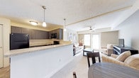 This single-floor residence features an open-concept floor plan that seamlessly connects the kitchen, dining, and living areas into one cohesive, functional space. The kitchen is outfitted with dark espresso cabinetry, black appliances, and a granite-look countertop peninsula that serves as both a workspace and a casual dining or entertaining surface. Pendant lighting above the peninsula adds a modern, polished touch, complementing the textured ceiling throughout.

Adjacent to the kitchen, the dining area accommodates a dark wood table and chairs, flowing naturally into the carpeted living room. The living room benefits from a ceiling fan for year-round comfort and is brightened by natural light streaming through large windows and glass patio doors. The patio doors provide both a visual connection to the outdoors and convenient exterior access.

The overall color palette is neutral and welcoming, with light-toned walls and carpeting that enhance the sense of openness. The layout is practical and well-proportioned, making the most of the available space while maintaining a comfortable, livable ambiance throughout.