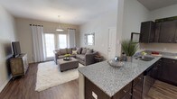 This single-floor residence offers a thoughtfully designed open-concept layout that seamlessly connects the kitchen and living areas, creating a welcoming and functional living environment. The kitchen features a generously sized granite-topped island with rich dark cabinetry, providing ample prep space and a natural gathering point. The surface's speckled grey-and-white tones lend a polished, contemporary feel to the space.

The adjoining living area is bright and airy, benefiting from an abundance of natural light streaming through large French doors dressed with white drape curtains. The neutral warm-grey walls and smooth ceilings contribute to the open, uncluttered atmosphere. A stylish pendant light anchors the space from above, while wood-look flooring runs throughout, adding warmth and continuity.

The living room is comfortably appointed with a sectional sofa, matching ottoman, and an accent mirror with a decorative frame that adds visual interest to the wall. A plush white area rug defines the seating zone, softening the overall aesthetic. The French doors open to the outdoors, enhancing the sense of space and allowing for easy indoor-outdoor flow. Overall, the residence strikes a balance between modern style and comfortable, everyday livability.