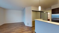 This single-floor space offers a clean, modern aesthetic defined by an open-concept layout and a neutral color palette. The interior is finished with warm hardwood flooring that runs throughout, complementing the crisp white walls and ceilings to create a light, airy atmosphere.

The kitchen area features a sleek peninsula with a white countertop, providing both a functional workspace and a natural divider between the kitchen and the living area. A stylish cylindrical pendant light hangs above, adding a contemporary touch to the space. A mirrored sliding door and built-in shelving are visible beyond the peninsula, offering practical storage solutions.

The open living area is generously proportioned, with a vaulted or angled ceiling detail that adds architectural interest and enhances the sense of vertical space. A floor vent ensures consistent climate control throughout. The layout flows naturally from the kitchen into the main living space, making the overall design feel connected and functional.

The absence of clutter and the predominantly white interior allow natural light to reflect easily, keeping the space feeling bright and welcoming. The combination of modern fixtures, durable flooring, and thoughtful spatial planning makes this a versatile and well-designed single-floor residence.