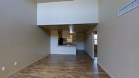 This single-story space makes a strong first impression with its dramatically high ceilings, which lend an airy, expansive quality to the interior. The open-concept layout connects the main living area to the kitchen seamlessly, promoting a sense of flow throughout the space. Warm, wood-style hardwood flooring runs consistently across the living area, contributing to a cohesive and inviting aesthetic.

The kitchen features a pass-through counter that opens directly to the living space, making it functional for both everyday use and entertaining. A pendant light hangs above the kitchen area, adding a subtle design detail, while a window fitted with horizontal blinds allows natural light to filter in. The kitchen is equipped with cabinetry and includes a refrigerator visible along the back wall.

A hallway extending from the main living area leads to additional rooms, with a doorway at the far end hinting at further living spaces. Natural light enters through windows along the right side of the space, brightening the neutral, white-toned walls. Overall, the space offers a clean, understated design with practical features throughout.